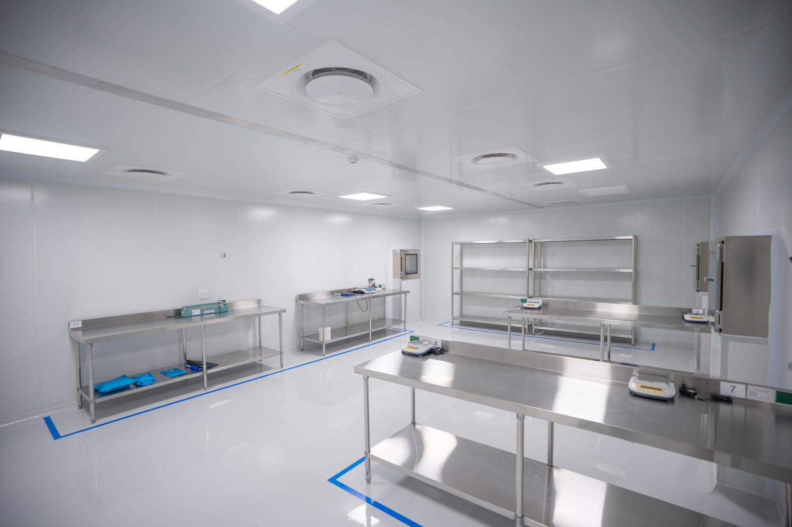 The GMP weighing and packaging room, stainless benches at numbered positions inside blue floor demarcation, pass-through hatches in the far wall